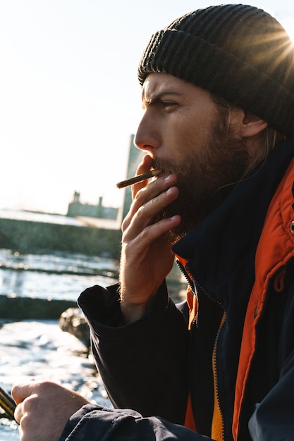 Imagen de un pescador joven apuesto con abrigo y sombrero en la orilla del mar fumando.