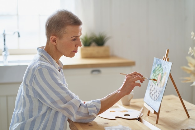 Imagen de pintura de mujer creativa de mediana edad en casa mientras está sentado en la mesa de madera en la cocina