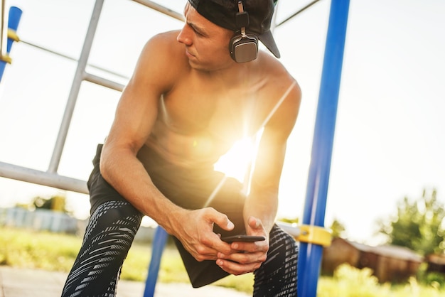 Imagen de primer plano del atleta joven musculoso relajante después de entrenar en el campo de entrenamiento al aire libre Guapo deportista sin camisa mirando hacia otro lado con el teléfono inteligente en las manos con auriculares escuchando música