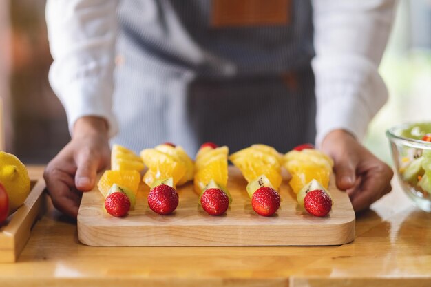 Imagen de primer plano de una cocinera sosteniendo y mostrando un plato de madera de frutas frescas mezcladas en brochetas en la cocina