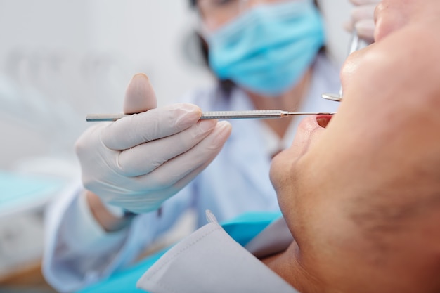 Imagen de primer plano de dentis usando un pequeño espejo y tallador al examinar los dientes de un paciente masculino