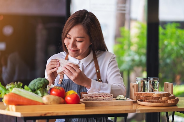 Imagen de primer plano de una hermosa cocinera cocinando y comiendo un sándwich de queso de jamón de trigo integral en la cocina