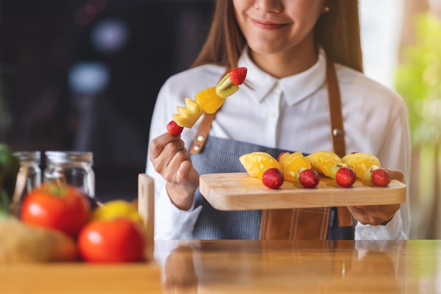 Imagen de primer plano de una hermosa cocinera sosteniendo frutas frescas mezcladas en brochetas en un plato de madera