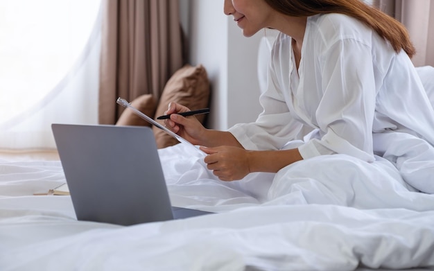 Imagen de primer plano de una hermosa joven asiática que usa una computadora portátil y trabaja en el papeleo mientras se sienta en la cama en casa