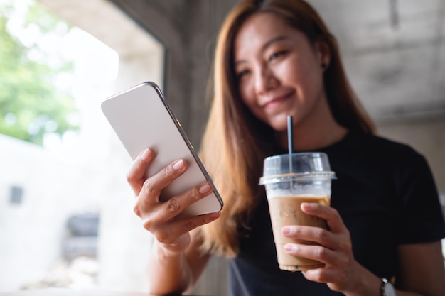 Imagen de primer plano de una hermosa joven asiática sosteniendo y usando un teléfono móvil mientras bebe café en la cafetería