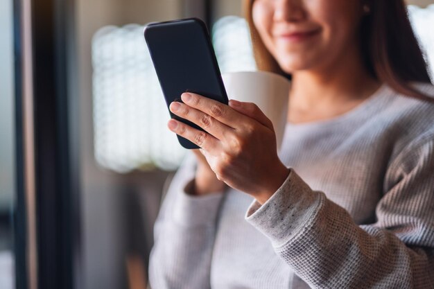 Imagen de primer plano de una hermosa joven asiática sosteniendo y usando un teléfono móvil mientras bebe café