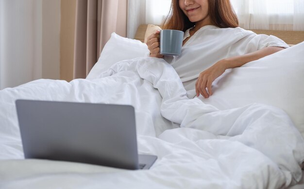 Imagen de primer plano de una hermosa joven asiática tomando café mientras mira una computadora portátil en una cama blanca y acogedora en casa