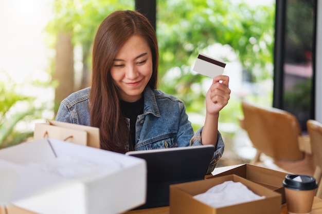 Imagen de primer plano de una joven asiática con tablet pc y tarjeta de crédito para compras en línea con caja de paquetería postal y bolsas de compras en la mesa