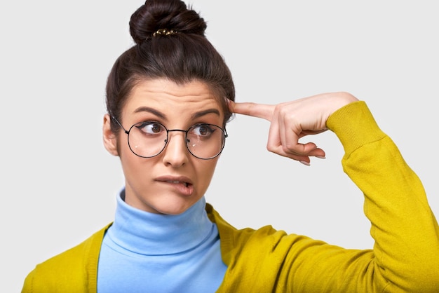 Imagen de primer plano de una joven incierta con un peinado de moño mordiéndose los labios siendo dudosa usando ropa casual y gafas mirando a un lado aislado sobre fondo blanco Confusión de emociones