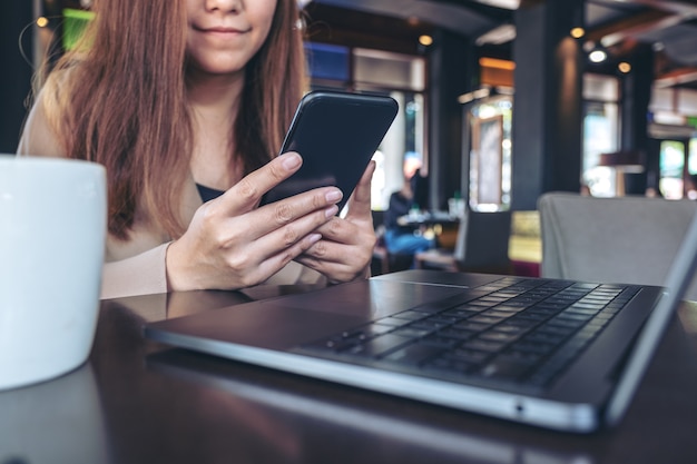 Imagen de primer plano de una mujer asiática sosteniendo, usando y mirando el teléfono inteligente mientras trabaja en la computadora portátil con una taza de café en la mesa en la oficina