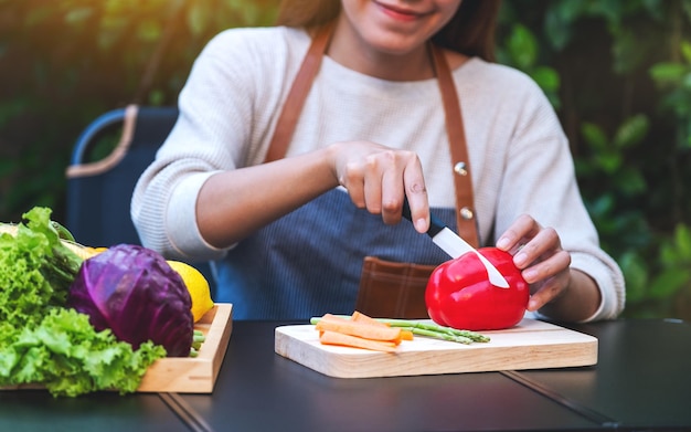 Imagen de primer plano de una mujer cortando y picando pimientos rojos con un cuchillo en una tabla de madera con verduras mixtas en una bandeja