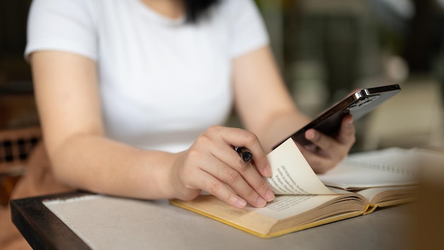 Imagen en primer plano de una mujer freelance usando su teléfono inteligente mientras lee un libro en una cafetería