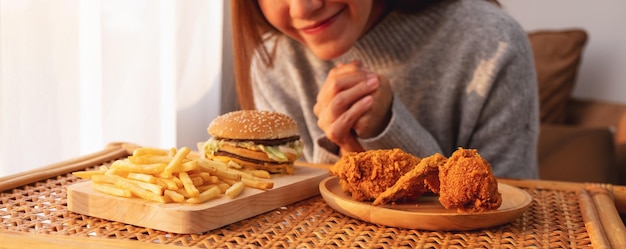 Imagen de primer plano de una mujer joven feliz de comer hamburguesas con papas fritas y pollo frito en la mesa en casa