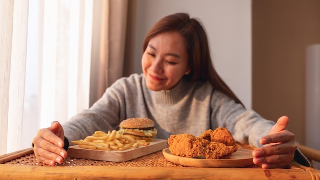 Imagen de primer plano de una mujer joven sosteniendo y comiendo una hamburguesa con papas fritas y pollo frito en la mesa en casa