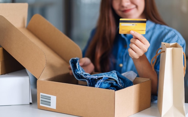 Imagen de primer plano de una mujer joven sosteniendo y usando tarjeta de crédito para compras en línea con bolsa de compras y caja de paquete postal de ropa sobre la mesa