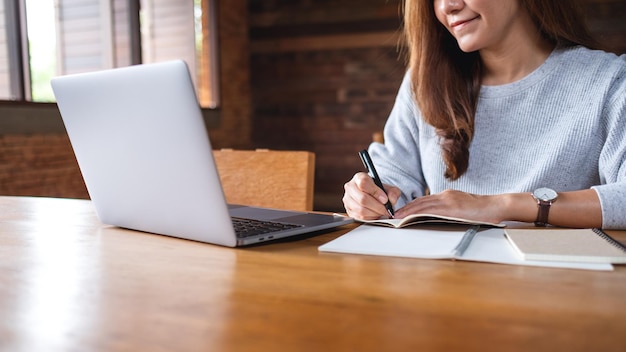 Imagen de primer plano de una mujer de negocios escribiendo en un cuaderno mientras trabaja en una computadora portátil en la oficina