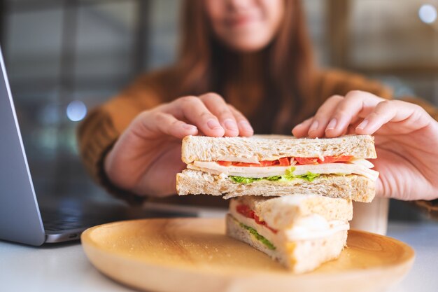 Imagen de primer plano de una mujer sosteniendo y comiendo sándwich de trigo integral mientras trabaja en la computadora portátil