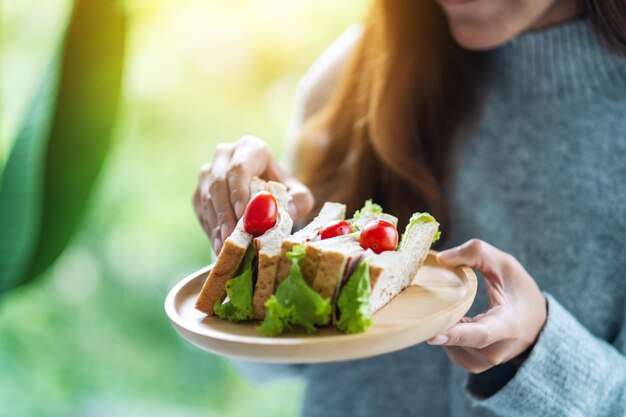 Imagen de primer plano de una mujer sosteniendo y comiendo un sándwich de trigo integral en un plato de madera