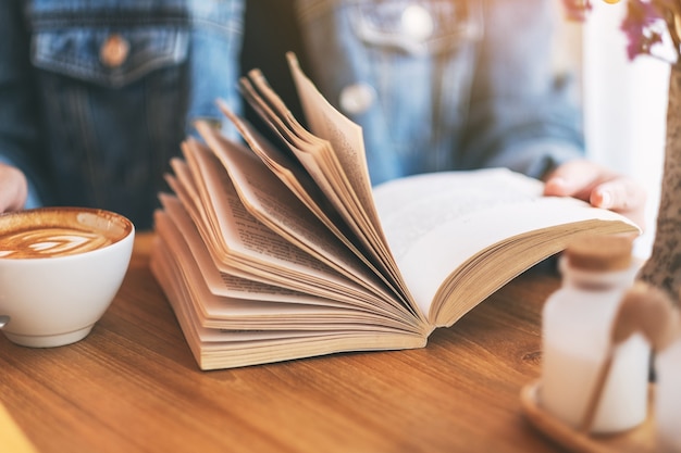 Imagen de primer plano de una mujer sosteniendo y leyendo un libro de novela vintage con taza de café en la mesa de madera