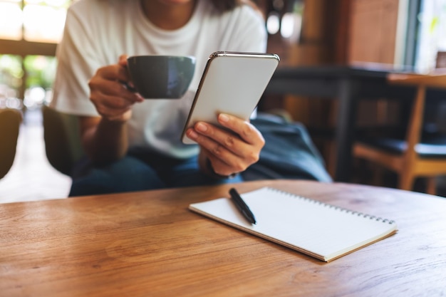 Imagen de primer plano de una mujer sosteniendo y usando un teléfono móvil mientras bebe café con un cuaderno en una mesa de madera