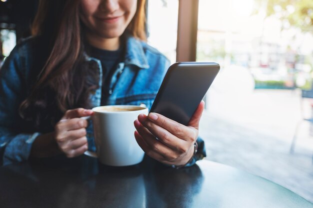 Imagen de primer plano de una mujer sosteniendo y usando el teléfono móvil mientras bebe café