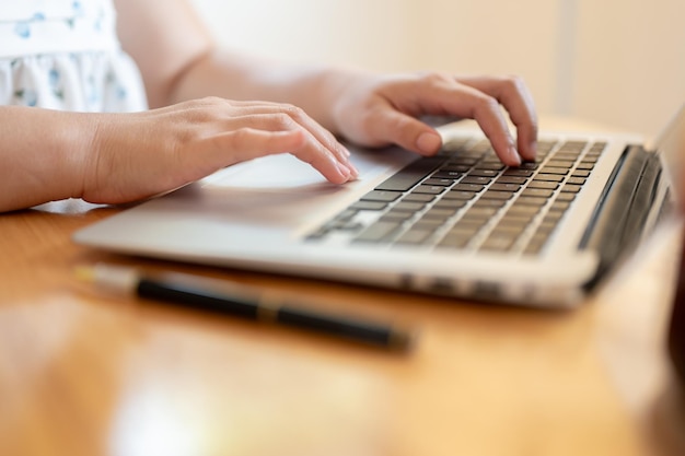Una imagen en primer plano de una mujer usando su portátil en una mesa en el interior escribiendo en el teclado de la computadora portátil