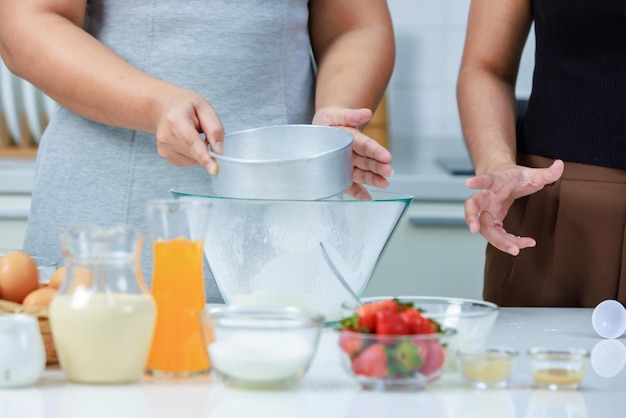 Foto imagen de primer plano de pasteleras de panadería femeninas irreconocibles, amas de casa que se mantienen de manos usando un tamiz inoxidable que tamiza la harina en polvo blanco en un cuenco de vidrio en una cocina completamente decorada con ingredientes para hornear.