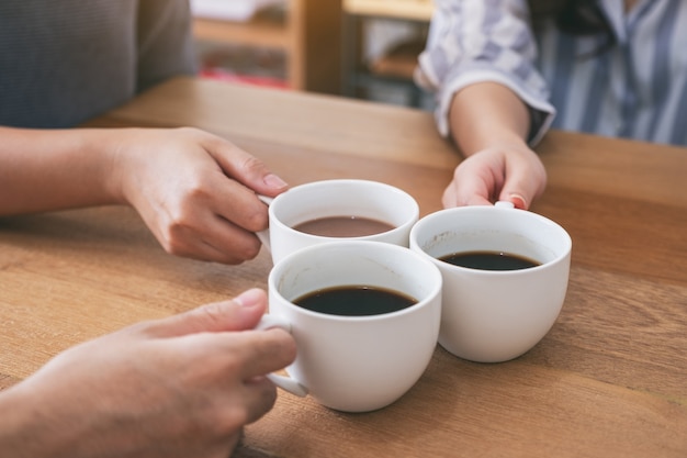 Imagen de primer plano de tres personas sosteniendo y tintineando tazas de café para beber en la mesa de madera