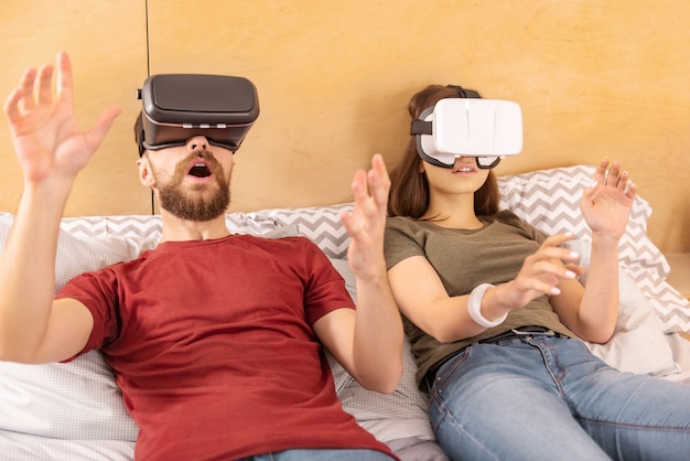 Imagen realista. Dulce pareja emocional sorprendida agitando las manos mientras está acostado y usando auriculares VR