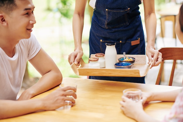 Imagen recortada de la camarera que sirve el desayuno que consiste en un delicioso yogur fresco y copos de maíz para una pareja joven