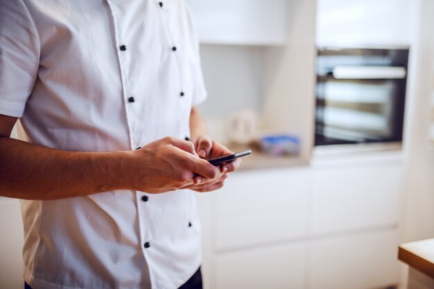 Imagen recortada del chef caucásico en uniforme de pie en la cocina y usando el teléfono inteligente.