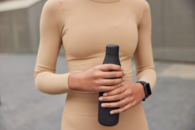 Imagen recortada de una deportista desconocida vestida con ropa activa beige que sostiene una botella de agua fresca y refrescante que usa poses de smartwatch al aire libre contra un fondo borroso Concepto de estilo de vida saludable