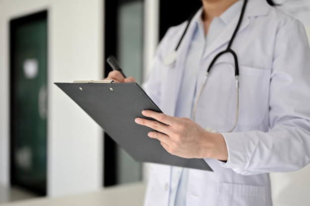 Imagen recortada de una doctora asiática profesional escribiendo algo en el portapapeles médico