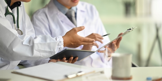 Imagen recortada de dos hombres inteligentes que trabajan como médico sosteniendo una tableta y un portapapeles mientras discuten y se sientan juntos en el escritorio del médico sobre el lugar de trabajo ordenado.