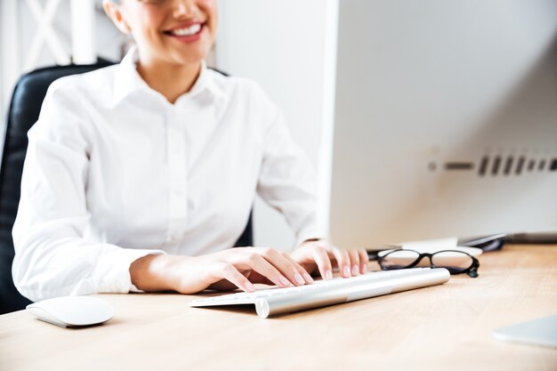 Imagen recortada de una empresaria feliz sonriente escribiendo en el teclado mientras está sentado en el escritorio de la oficina