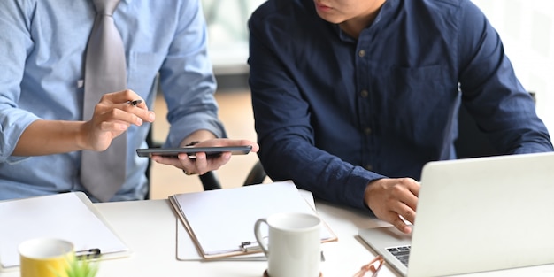 Imagen recortada del equipo de desarrolladores de negocios que trabaja junto con la computadora portátil y la tableta mientras está sentado en el escritorio de trabajo blanco que está rodeado por el portapapeles y la taza de café, concepto de colaboración.