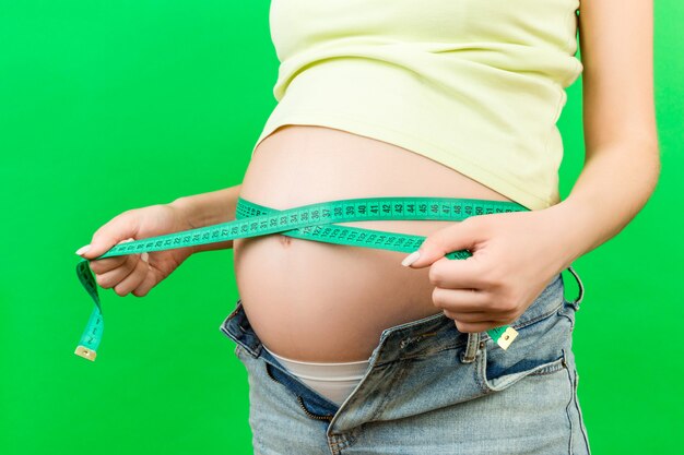 Imagen recortada de la futura madre en jeans desabrochados midiendo su vientre embarazado con una cinta métrica en colores de fondo. medición de entímetro
