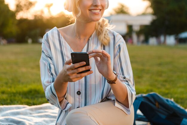 Imagen recortada de una hermosa joven rubia relajándose en un césped en el parque, escuchando música con auriculares mientras usa el teléfono móvil