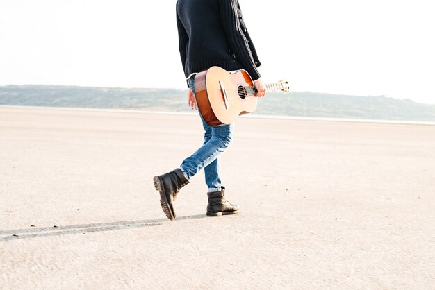 Imagen recortada de un hombre casual sosteniendo la guitarra y caminando por la orilla del mar