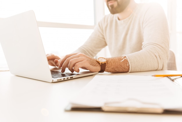 Imagen recortada del hombre caucásico con reloj de pulsera y ropa casual trabajando en la computadora portátil, mientras está sentado en la mesa interior