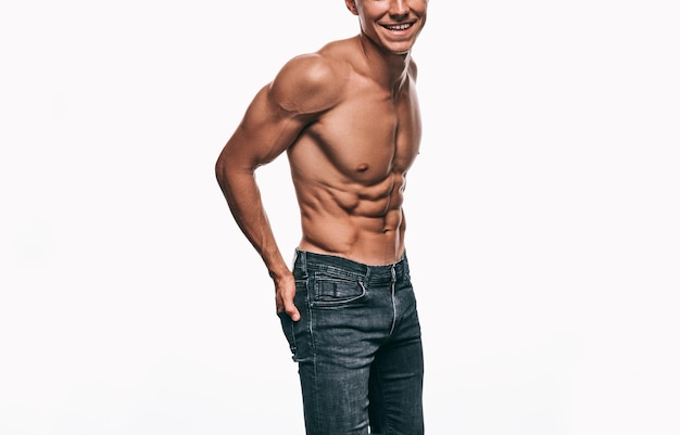 Imagen recortada de hombre joven sonriendo y usando jeans mostrando su cuerpo sexy aislado en la pared blanca Hombre culturista sin camisa con abdomen sexy posando sobre la pared blanca Concepto de gente de deporte de salud