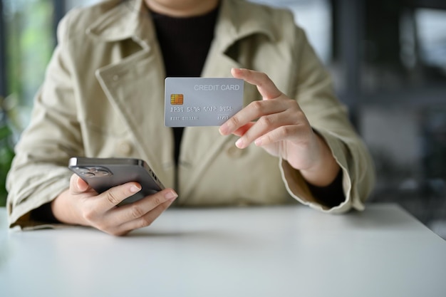 Imagen recortada de una joven asiática que sostiene una tarjeta de crédito y un teléfono inteligente en su escritorio