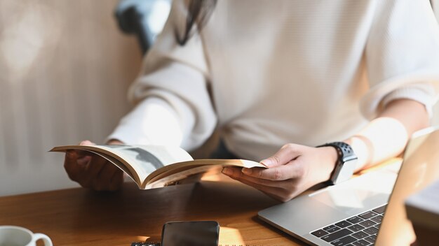 Imagen recortada de la joven y bella mujer sosteniendo / leyendo un libro en sus manos frente a una computadora portátil, una taza de café y un teléfono inteligente en la moderna mesa de madera con una cómoda sala de estar como