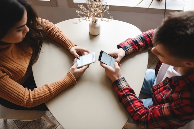 Imagen recortada de una linda pareja amorosa joven sentada en un café en el interior usando teléfonos móviles.