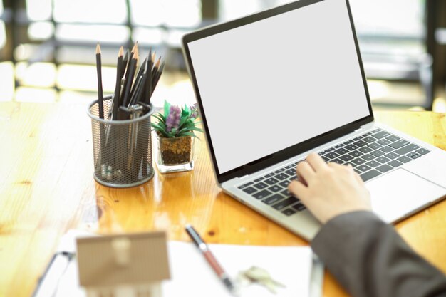 Imagen recortada de un lugar de trabajo con maqueta de pantalla en blanco de computadora portátil y una mano femenina escribiendo en un teclado