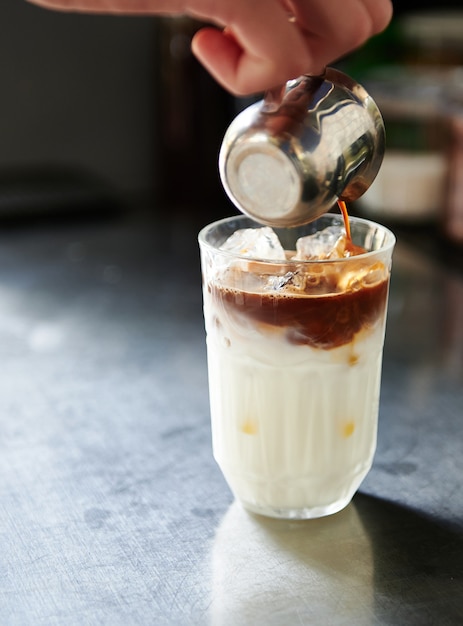 Imagen recortada de la mano del barista vertiendo café en un vaso con leche batida con hielo mientras prepara un café con leche helado