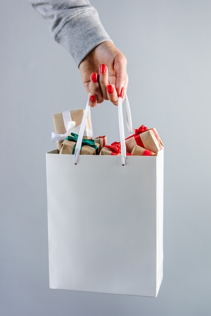 Foto imagen recortada de mano femenina con uñas pulidas rojas sosteniendo una bolsa de compras blanca lisa llena de cajas de regalo de navidad