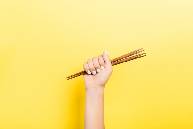 Imagen recortada de la mano femenina que sostiene los palillos en el puño sobre fondo amarillo. Concepto de comida asiática con espacio de copia