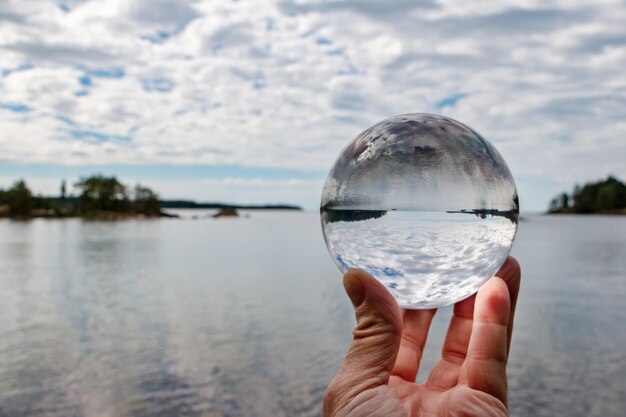 Imagen recortada de la mano sosteniendo una bola de cristal contra el lago y el cielo nublado