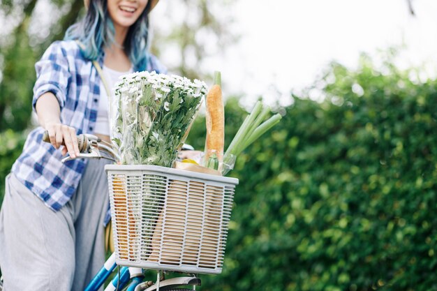 Imagen recortada de una mujer asiática joven emocionada montando bicicleta en el parque con una cesta de flores y comestibles